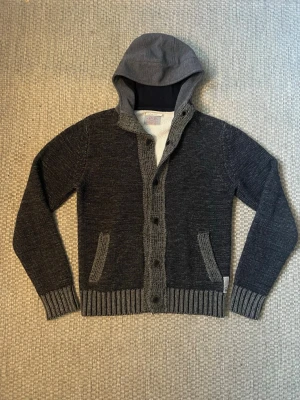 Mörkgrå cardigan jacka från Replay - Snygg mörkgrå cardigan jacka från Replay med stickade detaljer och huva i sweatshirt-tyg. Jackan har knappar framtill, ribbade muddar och två fickor. Perfekt för dig som gillar en chill och avslappnad stil.