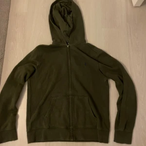 Olivgrön hoodie från Polo Ralph Lauren - Olivgrön hoodie från Polo Ralph Lauren med dragkedja och klassisk broderad logga på bröstet. Tröjan har huva, känguruficka och är tillverkad i mjuk bomullsmix. Perfekt för chill dagar och snygg streetstyle.L 14-16 passar mellan 150-164 cm