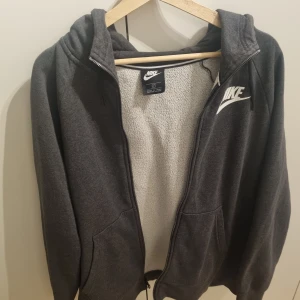 Mörkgrå hoodie från Nike - Mörkgrå hoodie från Nike med dragkedja och vit logga på bröstet. 