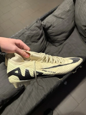 Nike Mercurial AirZoom Academy fotbollsskor - Säljer ett par Nike Mercurial AirZoom Academy ag/fgfotbollsskor i ljusbeige med svarta detaljer och logga. Skorna har snörning, platt sula med dobbar FG/ag  och en rund tå. Materialet är syntet och designen är riktigt sportig, perfekt för dig som vill ha grymma skor på planen. Använda under 1 match och en träning på konstgräs. Säljs för 250kr.