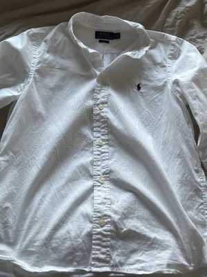 Vit skjorta Polo Ralph Lauren Slim - Klassisk vit skjorta från Polo Ralph Lauren i slim fit-modell. Skjortan har långa ärmar, knappar framtill och den ikoniska broderade loggan på bröstet. Tillverkad i mjuk bomull och passar perfekt till en clean och stilren look.