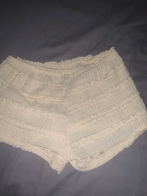 Pastel gul volangshorts med spetsdetaljer - Säljer ett par pastel gula shorts med volang och spetsdetaljer. De har en loose passform och är tillverkade i ett mjukt, strukturerat tyg som känns luftigt. Perfekta för varma dagar och har en söt, feminin look med fina detaljer längs kanterna.