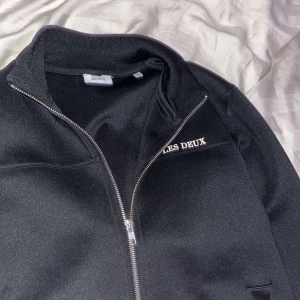 Svart bomberjacka från Les Deux - Svart bomberjacka från Les Deux med stilren design och silverfärgad dragkedja framtill. Jackan har ståkrage, två sidofickor och broderad logga på bröstet. Perfekt för dig som gillar enkel och clean stil.