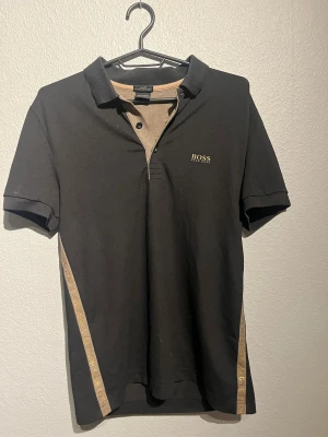 Svart slim fit piké från Hugo Boss - Svart pikétröja från Hugo Boss med beige detaljer och logga på bröstet. Slim fit-modell med korta ärmar och knappslå framtill. Snygg beige kant med Hugo Boss-tryck längs sidorna. Perfekt för dig som gillar stilrena och exklusiva plagg. Pris kan diskuteras bara hör av dig!