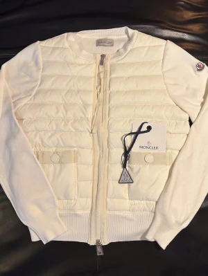 Moncler Cardigan  - Säljer en fräsch vit cardigan jacka från Moncler med quiltad front och stickade ärmar. Jackan har dragkedja, två fickor med knappstängning och Moncler-logga på ärmen. Perfekt för dig som vill ha en clean och stilren look.