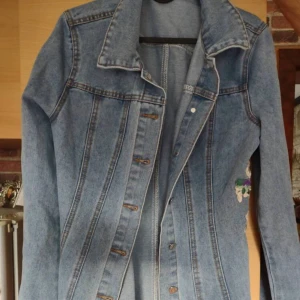 Blå jeansklänning med broderade blommor - Säljer en lång jeansklänning i ljusblå denim med snygga broderade blommor i rött, lila, grönt och vitt på sidan. Klänningen har knappar framtill och klassisk krage. Perfekt för dig som gillar detaljer och vill sticka ut.