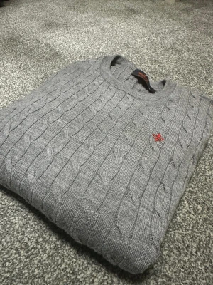 Morris tröja - Snygg grå kabelstickad tröja från Stockholm Morris med rund hals och ett litet rött broderat emblem på bröstet. Tröjan har ett klassiskt mönster och är tillverkad i mjukt stickat material som passar perfekt när du vill ha en chill och stilren look.