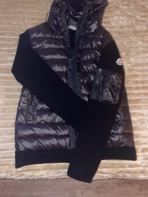 Svart Moncler pufferjacka med huva - Säljer en svart pufferjacka från Moncler med glansig yta och stickade svarta ärmar. Jackan har huva, dragkedja framtill och en ficka med dragkedja på ärmen där Moncler-loggan sitter. Perfekt för dig som gillar stilren och en lite skönare look. Den här inga defekter och fraktar direkt 😃