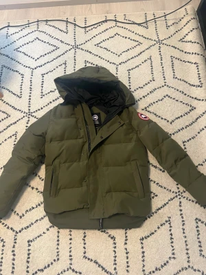 Canada Goose MacMillan army green xs - Säljer en olivgrön dunjacka från Canada Goose i storlek XS. Jackan har huva, två stora fickor framtill och klassisk patch på ärmen. Insidan är svart och jackan stängs med både dragkedja och knappar. Perfekt för kalla dagar och riktigt snygg streetstil.