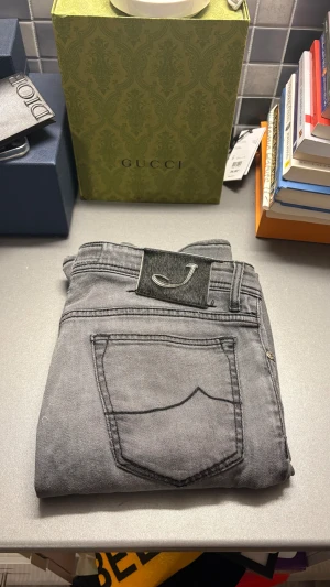 Grå smala jeans från Jacob Cohen - Säljer ett par grå jeans från Jacob Cohen med snyggt J-emblem på bakfickan. Modellen är smal och har klassisk femficksdesign, med svarta sömmar och knappgylf. Jeansen är tillverkade i mjukt bomullsmaterial med stretch för extra komfort.