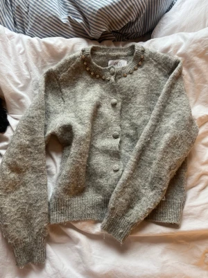 Grå kofta med pärldetaljer från Zara - Mysig grå kofta från Zara med rund hals och dekorativa pärlor längs kragen. Koftan är stickad i mjukt material och har ribbade muddar samt knappar framtill. Perfekt för lager-på-lager under kyliga dagar.