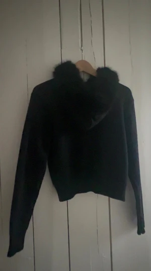 Svart cardigan jacka med fuskpäls - Svart cardigan jacka med fluffig fuskpäls runt kragen och huvan. Jackan har lång ärm, ribbad nederkant och dragkedja framtill. Perfekt för dig som vill ha en mysig och stilren look med lite extra edge. Ifrån Gina14+ köpte den för 399 o aldrig använt den men tagit av lappen, storlek M men är liten i storlek så skulle säga S