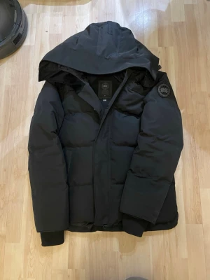 Svart pufferjacka från Canada Goose - Svart pufferjacka från Canada Goose med stor huva och diskret logotyp på ärmen. Jackan har quiltad design, dragkedja och ribbade muddar vid ärmslut. Perfekt för kalla dagar och har en clean, stilren look som funkar till streetwear.