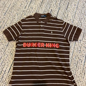 Brun randig polotopp från Ralph Lauren - Säljer en brun och vitrandig polotopp från Ralph Lauren i storlek XXL. Toppen har klassisk krage, korta ärmar och knäppning med två knappar framtill. Materialet är 100% bomull och på bröstet finns Ralph Laurens logga samt ett tryck med 'BURGER KING' i rött. Jag köpte en polo tröjä från Tise x Burger King popup! 