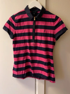 Randig polotopp från Tommy Hilfiger - Säljer en snygg polotopp från Tommy Hilfiger med breda rosa och marinblå ränder. Toppen har korta ärmar, krage och en guldfärgad dragkedja framtill. Liten logga på bröstet. Perfekt för dig som gillar sportig och färgstark stil.
