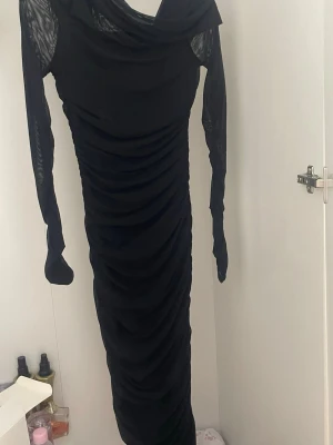 Svart långklänning med drapering - Snygg svart långklänning med draperade detaljer och transparenta långärmar. Klänningen har en cool omlottkrage och är figurnära med ett elegant fall. Perfekt för dig som gillar stilrena och trendiga plagg med lite edge.