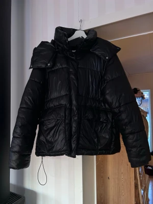Svart pufferjacka med dragkedja - Säljer en svart pufferjacka med hög krage och stora fickor framtill. Jackan har både dragkedja och tryckknappar, samt en quiltad design som ger extra volym. Materialet är glansigt och känns syntetiskt, perfekt för kalla dagar. Fint skick!!