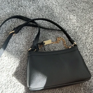 Svart axelväska med guldkedja - Snygg svart axelväska i skinn med en elegant guldkedja som detalj. Perfekt för både vardag och fest! Väskan har en justerbar axelrem och en dragkedja för säker förvaring.