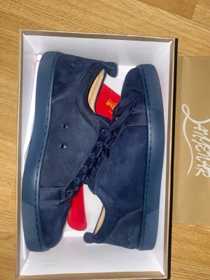 Mörkblå sneakers från Christian Louboutin - Säljer ett par mörkblå sneakers från Christian Louboutin med klassisk röd sula och snygg signatur på undersidan. Skorna är i mocka med rund tå, platt sula och snörning. Perfekt för dig som gillar exklusiva och stilrena sneakers med lyxig känsla. Samt är dem helt nya från boxen