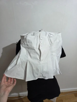 Vit plisserad kortkjol från H&M - Säljer en vit plisserad kortkjol från H&M med bred linning och tre dekorativa knappar framtill. Kjolen har en klassisk och fräsch look, perfekt för dig som gillar stilrena outfits. Materialet är bomull och kjolen har en normal passform.