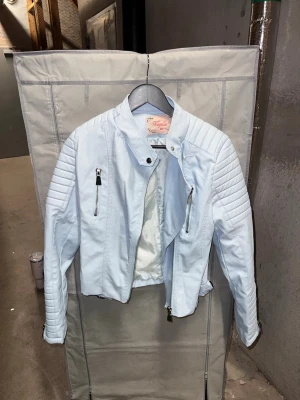 Ljusblå skinnjacka från Voyelles - Säljer en ljusblå skinnjacka från Voyelles med coola detaljer som dragkedjor framtill, quiltade partier på axlar och ärmar samt snörning i nederkant bak. Jackan har en kort, figurnära passform och ståkrage med tryckknapp. Perfekt för dig som vill sticka ut!