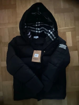 Svart pufferjacka från Burberry - Säljer en svart pufferjacka från Burberry med avtagbara ärmar så du kan göra om den till väst. Jackan har huva med klassiskt rutigt foder och tydlig Burberry-logga på ärmen. Perfekt för kalla dagar och riktigt snygg streetstil.