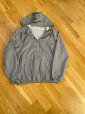 Grå vindjacka med huva - Säljer en stilren grå vindjacka med huva och dragkedja framtill. Jackan har elastiska muddar vid ärmslut och nederkant, samt två sidofickor. Perfekt för blåsiga dagar och enkel att slänga på över en hoodie.