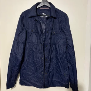 Blå vindjacka från Snoot Stockholm - Snygg marinblå vindjacka från Snoot Stockholm med klassisk krage och tryckknappar framtill. Jackan har två bröstfickor med knappstängning och är tillverkad i lätt, vattenavvisande syntetmaterial. Perfekt för dig som gillar stilren och funktionell design.