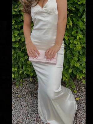 Champagnefärgad långklänning Nelly - Elegant långklänning i champagnefärgad satin från Nelly. Klänningen har smala axelband, öppen rygg med snörning och figurnära passform. Materialet är glansigt och mjukt, vilket ger en lyxig känsla. Perfekt för speciella tillfällen. Klänningen är endast andvänd en gång och skicket är som nytt. Den har inga defekter. 