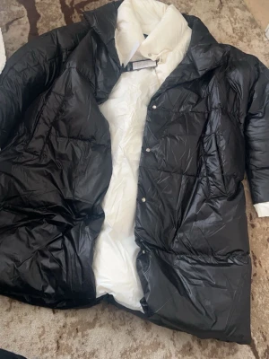 Svart och vit lång dunjacka Hollies - Säljer en lång dunjacka från Hollies i svart med vit insida. Jackan har en quiltad design, stora fickor och knappar framtill. Perfekt för kalla dagar med sin fluffiga och varma känsla. Stilig och modern look som sticker ut.