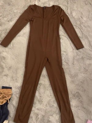 Brun långärmad jumpsuit - Säljer en brun jumpsuit med lång ärm och rundad halsringning. Modellen är figurnära och har långa ben, perfekt för dig som gillar en enkel och stilren look. Materialet känns mjukt och stretchigt, vilket gör den bekväm att bära hela dagen. Kan eventuellt tänka mig gå ner i pris