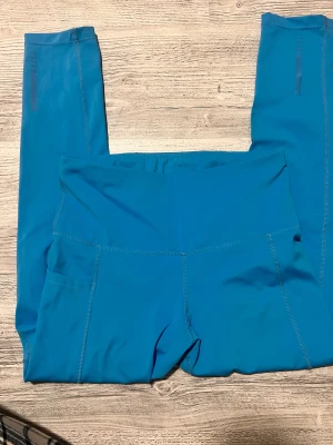 Blå höga leggings med fickor - Snygga blå leggings med hög midja och praktiska sidofickor. Tillverkade i ett stretchigt syntetmaterial som passar perfekt för träning eller chill. Söm-detaljer längs benen och diskreta mönster vid vaden ger extra stilpoäng.