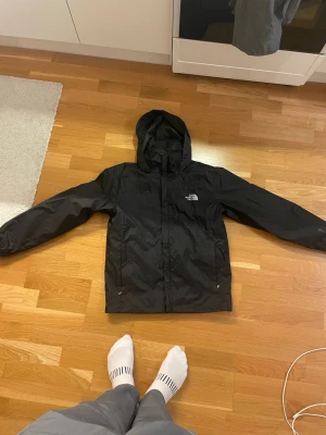 Svart vindjacka från The North Face - Svart vindjacka från The North Face Summit Series med huva och dragkedja framtill. Storlek L men sitter mer som en medium. Jackan har två sidofickor med dragkedja och elastiska muddar vid ärmsluten. Materialet är lätt och syntetiskt, perfekt för blåsiga dagar. Använd 1 gång