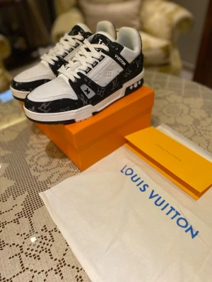 Louis Vuitton svartvita sneakers - Louis Vuitton sneakers i svart och vitt med klassiskt monogrammönster. Skorna har snörning, rund tå och platt sula. Materialet är en mix av skinn och textil, med tydliga LV-loggor på både ovansida och sula. Snygg kontrast mellan svart och vit, perfekt för dig som gillar lyxiga detaljer.
