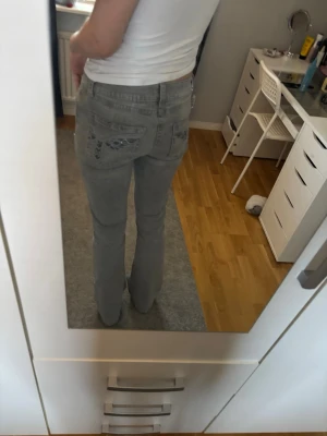Grå bootcut jeans med broderade fickor - Säljer ett par grå jeans med bootcut-snitt och låg midja/mid waist. Baksidan har snygga broderade detaljer på fickorna som ger extra stilpoäng. Jeansen är tillverkade i klassiskt denim och har en riktigt chill vibe. Perfekta för dig som vill ha något som sticker ut lite. Har använt 2 gånger!💞
