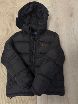 Svart pufferjacka från Polo Ralph Lauren - Svart pufferjacka från Polo Ralph Lauren med huva och röd broderad logga på bröstet. Jackan har dragkedja framtill och två sidofickor med dragkedja. Perfekt för kalla dagar och har en schysst, lite puffig look.