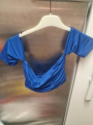 Blå topp - Säljer en blå topp och korta ärmar. Toppen har snygga rynkade detaljer framtill och är tillverkad i ett mjukt syntetmaterial. Perfekt för dig som gillar trendiga och färgstarka plagg.