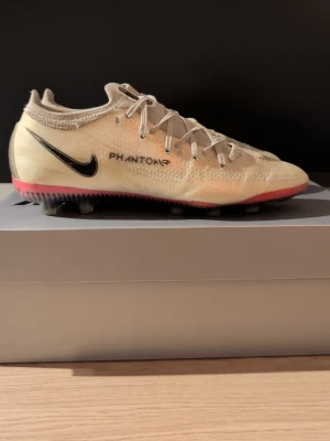 Nike Phantom Elite fotbollsskor - Säljer ett par Nike Phantom Elite fotbollsskor i beige med svarta detaljer och rosa sula. Skorna har snörning, rund tå och är tillverkade i syntetmaterial med stickad krage för extra komfort. Perfekta för dig som vill ha grym touch och kontroll på planen.