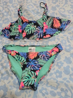 Färgglad bikini från H&M - Säljer en bikini från H&M med tropiskt bladmönster i neonfärger som blå, grön, rosa och orange mot svart bakgrund. Toppen har smala axelband och volangdetalj, och trosan är klassisk med mintgrön insida. Perfekt för strandhäng! Passar bra för 8 årig tjej. 