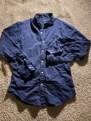 Marinblå skjorta från Polo Ralph Lauren - Snygg marinblå skjorta från Polo Ralph Lauren i slim fit. Skjortan har klassisk krage, vita knappar och det ikoniska broderade logot på bröstet. Tillverkad i mjuk bomull och har långa ärmar. Perfekt för dig som gillar stilrena och tidlösa plagg.
