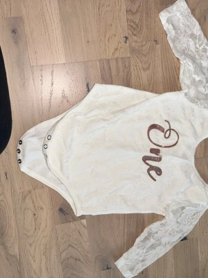 Vit spetsbody med glittertext Vit body - Säljer en vit body med långa spetsärmar och tryckknappar i grenen. Framsidan har glittrig text med ordet 'One' i rosa. Materialet är en kombination av spets och mjuk bomull på insidan, vilket gör den bekväm och snygg. Perfekt för speciella tillfällen. För 1år