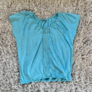 Ljusblå blus med spets från Janina - Ljusblå blus från Janina med korta ärmar och rund halsringning. Framsidan har en bred spetsdetalj som ger en söt touch. Blusen är tillverkad i ett lätt och mjukt bomullsmaterial, perfekt för varmare dagar. Den är stretchig. Storlek EUR 42