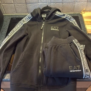 Svart EA7 hoodie med logga och dragkedja - Svart hoodie från EA7 Emporio Armani med dragkedja framtill och huva. Snygga loggoband längs ärmarna och EA7-tryck på bröstet. 