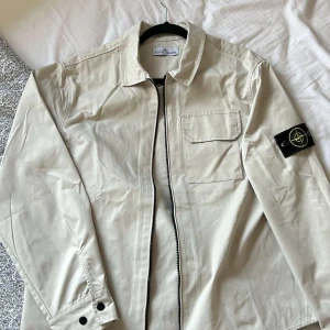 Beige overshirt från Stone Island - Snygg beige overshirt från Stone Island med klassisk krage, bröstficka och ikonisk patch på ärmen. Jackan har dragkedja framtill och är tillverkad i bomull, perfekt för lager-på-lager-stil. Passar dig som gillar clean och stilren look.