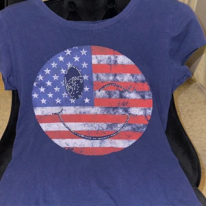 Blå t-shirt med amerikanskt smileytryck - Blå t-shirt med ett stort tryck av en smiley i amerikanska flaggans färger på bröstet. Trycket har vita stjärnor och röda ränder, samt glittriga detaljer. 