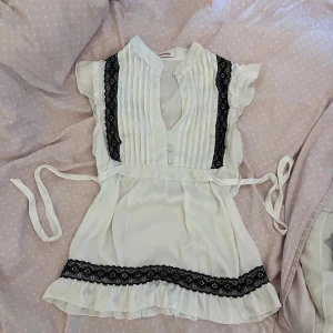 Vit blus med svarta spetsdetaljer - Supersöt vit blus med svarta spetsdetaljer längs sidorna och nertill. Blusen har knappar framtill, volangärmar och knytband i midjan. Plisserad upptill och lätt v-ringad för en feminin look.