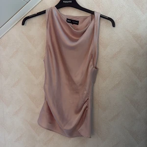 Ljusrosa satinlinne från Zara - Säljer ett ljusrosa/smutsrosa linne från Zara i glansig satin. Toppen har en snygg draperad halsringning och är ärmlös med en lätt loose passform. Perfekt att styla med jeans eller kjol för en trendig look. Den är i bra skick men har en enstaka repning (sista bilden) men tycker inte de är något man tänker på