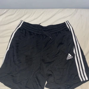Svarta Adidas shorts med vita ränder - Svarta shorts från Adidas med klassiska vita ränder längs sidorna och Adidas-logga på vänster ben. De har elastisk midja med snörning för skön passform. Perfekta för träning eller chill. Materialet känns mjukt och lätt, troligen bomull eller polyester.