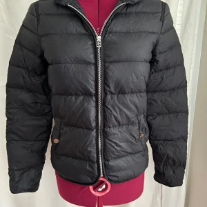 Svart dunjacka från InWear, stl 34 - Snygg svart dunjacka från InWear i storlek 34. Jackan är quiltad med horisontella sömmar, har dragkedja framtill och två fickor med tryckknapp. På insidan av kragen syns tecken på användning (se bild 5)