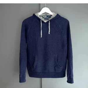 Mörkblå kashmir hoodie Polo Ralph Lauren - Exklusiv mörkblå hoodie från Polo Ralph Lauren i 100% kashmir. Tröjan har grå insida på huvan, vita snören och klassisk känguruficka framtill. Långärmad och supermjuk, perfekt för dig som gillar stilren och lyxig streetwear.